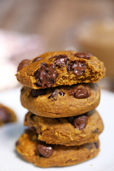 4 stacked pumpkin chocolate chip cookies with a bite out of the top cookie on a white counter.
