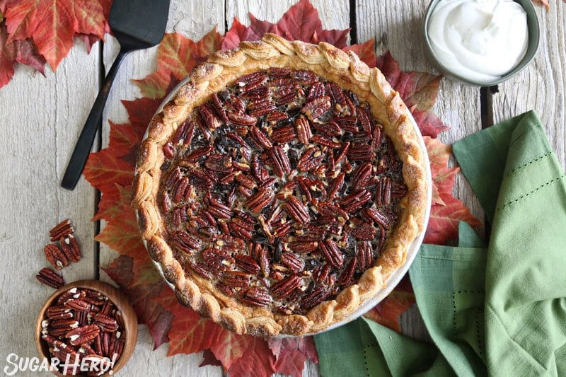 Deep Dish Pecan Pie - a gloriously gooey pie, packed with toasted pecans and chunks of chocolate. For serious pie lovers only! | From SugarHero.com Deep Dish Pecan Pie - overhead picture of whole pie with a bowl of whipped cream and pecans around the pie | From SugarHero.com