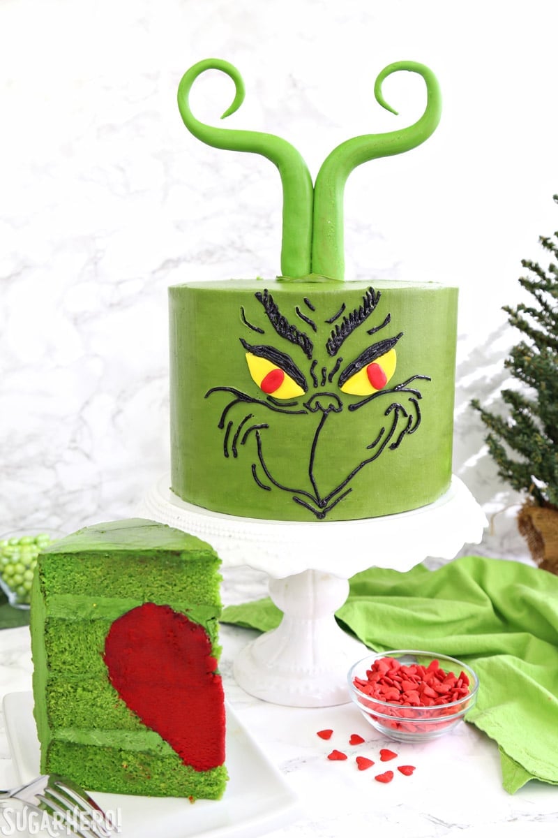 Grinch Cake - front of Grinch cake on a cake stand, with a slice of cake showing the red heart inside | From SugarHero.com