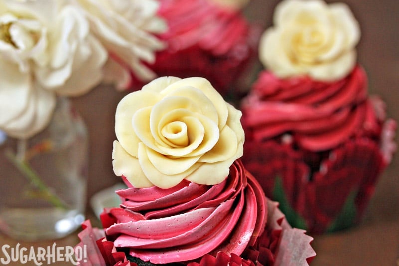 Chocolate Rose Cupcakes are moist chocolate cupcakes, topped with a beautiful swirl of red frosting and a sweet white chocolate rose. Learn how to make these hand-formed edible roses, and use them on cupcakes, cakes, and so many more desserts! | From SugarHero.com Chocolate Rose Cupcakes - The top of a cupcake to display the chocolate rose on top. | From SugarHero.com