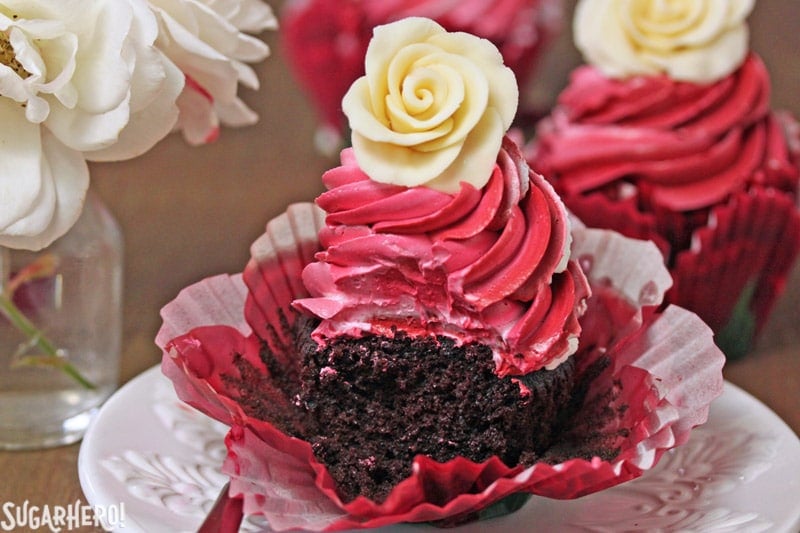 Chocolate Rose Cupcakes are moist chocolate cupcakes, topped with a beautiful swirl of red frosting and a sweet white chocolate rose. Learn how to make these hand-formed edible roses, and use them on cupcakes, cakes, and so many more desserts! | From SugarHero.com Chocolate Rose Cupcakes - A single cupcake with a bite taken out of it. | From SugarHero.com