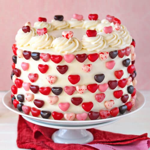 Pink and red velvet cake on a white cake stand, with red candy heart decorations.