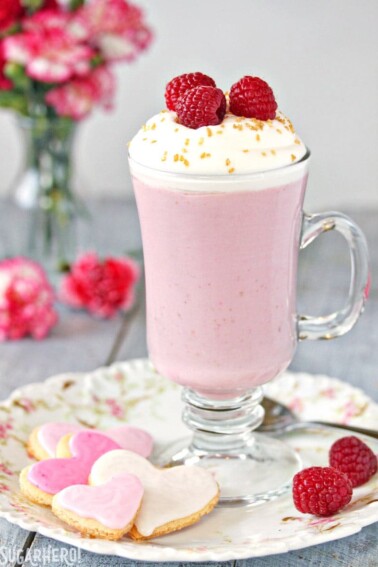 A mug of Raspberry White Hot Chocolate on a floral plate next to cookies and berries.