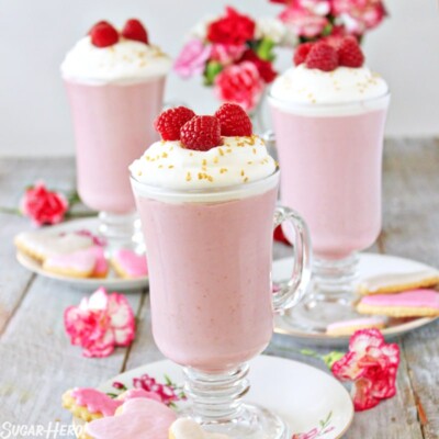 3 mugs of Raspberry White Hot Chocolate on floral plates next to cookies and flowers.