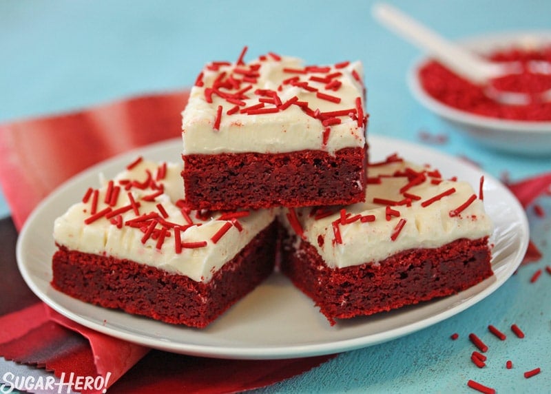 These Red Velvet Bars have the great taste of red velvet cake, in a portable cookie bar! | From SugarHero.com Red Velvet Bars - Three separate Red Velvet Bars on a plate with red homemade sprinkles. | From SugarHero.com