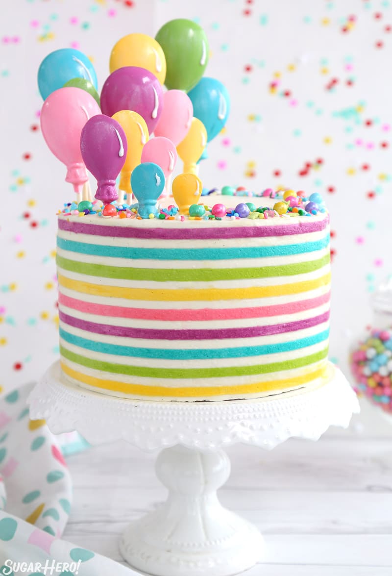 Looking for the perfect party cake?! Buttercream stripes are trendy right now, and they add a bit of fun to any cake design. Pair them with candy balloons for the ultimate birthday cake! | From SugarHero.com Striped Buttercream Balloon Cake on a white cake stand in front of a polka dot background.