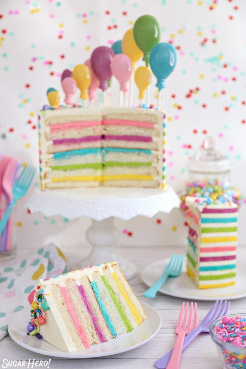 Striped Buttercream Balloon Cake sliced open on a white cake stand, with several cake slices on white plates in front of it.