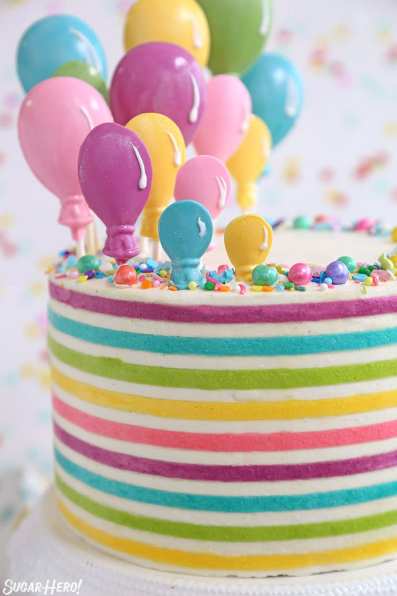 Looking for the perfect party cake?! Buttercream stripes are trendy right now, and they add a bit of fun to any cake design. Pair them with candy balloons for the ultimate birthday cake! | From SugarHero.com Striped Buttercream Balloon Cake on a white cake stand in front of a polka dot background.