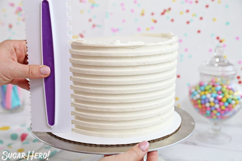 Looking for the perfect party cake?! Buttercream stripes are trendy right now, and they add a bit of fun to any cake design. Pair them with candy balloons for the ultimate birthday cake! | From SugarHero.com Hand holding a cake scraper creating white stripes on a layer cake.