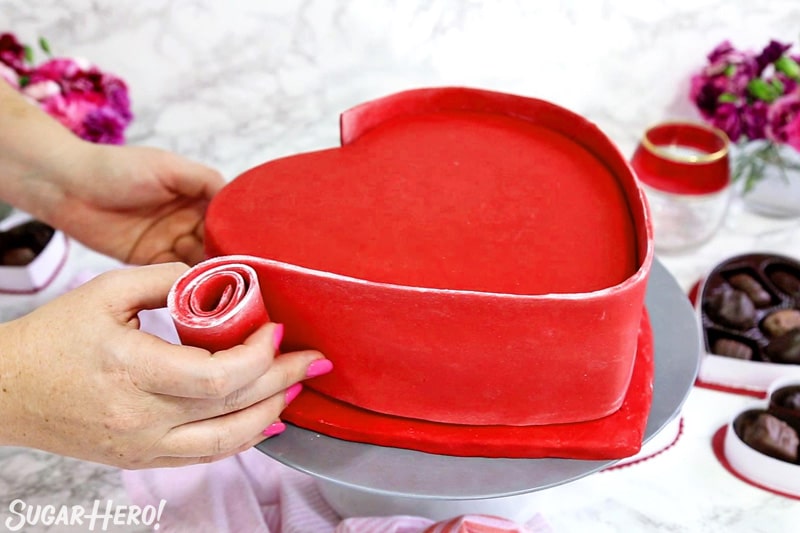 This Box of Chocolates Cake is two treats in one—a chocolate cake filled with strawberry buttercream, plus gourmet chocolates on top! Great for Valentine's Day! | From SugarHero.com Box of Chocolates Cake - unrolling the red fondant panel along the sides of the cake | From SugarHero.com
