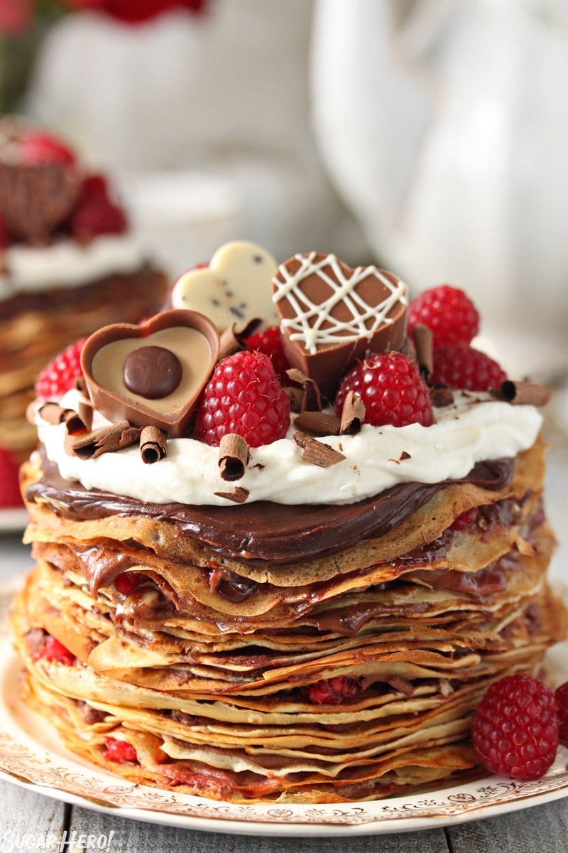 These Chocolate Raspberry Mini Crepe Cakes are so beautiful and elegant! Miniature crepes are layered with chocolate and fresh raspberries to make a unique and delicious dessert. | From SugarHero.com Chocolate Raspberry Mini Crepe Cakes - A single cake with heart shaped chocolates and raspberries on top. | From SugarHero.com