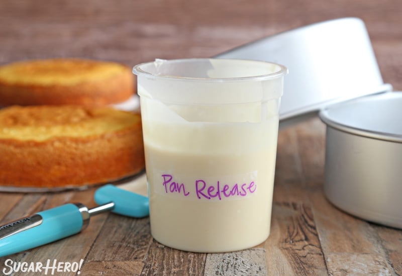 Container of homemade pan release with a pastry brush, silver cake pans, and cooked yellow cakes in the background