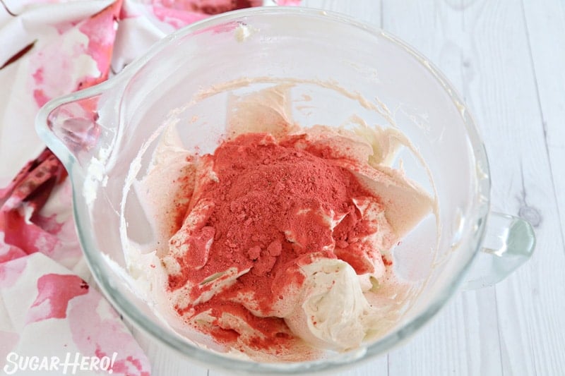 This Strawberry Buttercream is incredible! With a smooth, creamy texture and bright fresh strawberry flavor, it's going to be your new favorite frosting to put on everything! | From SugarHero.com Strawberry Buttercream Frosting - freeze-dried strawberry powder scattered on top of white frosting in bowl | From SugarHero.com