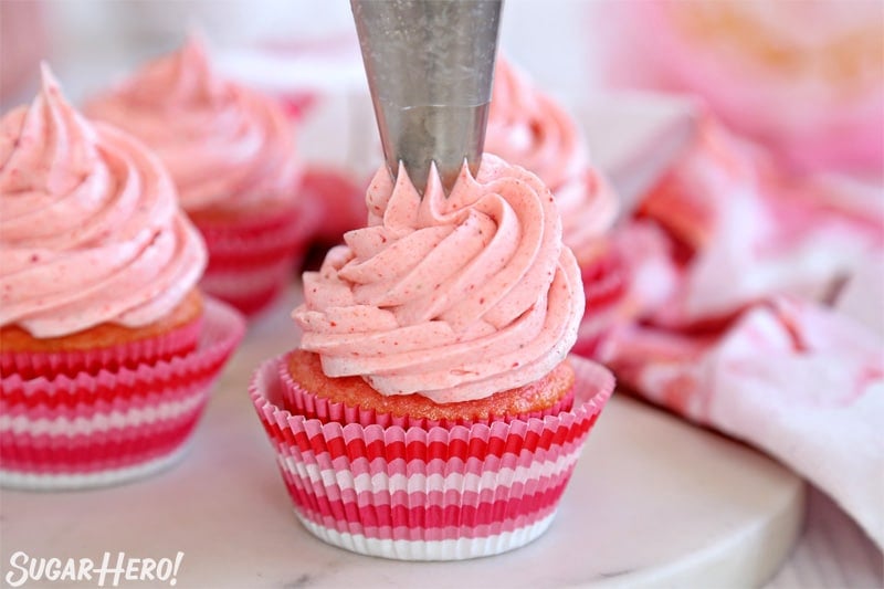 This Strawberry Buttercream is incredible! With a smooth, creamy texture and bright fresh strawberry flavor, it's going to be your new favorite frosting to put on everything! | From SugarHero.com Strawberry Buttercream Frosting - piping a big swirl of pink frosting on top of a cupcake | From SugarHero.com