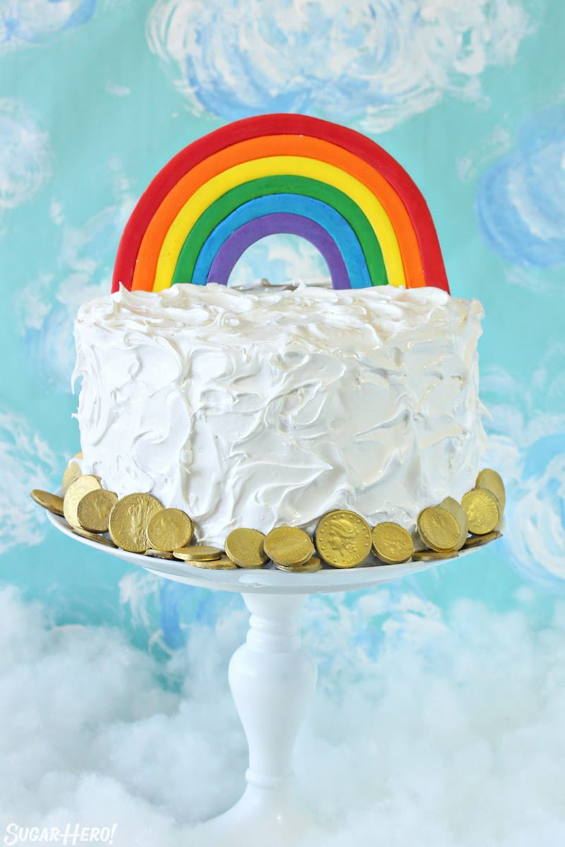 Cake with fluffy white frosting and a rainbow cake topper on a white cake stand with a blue cloud background.