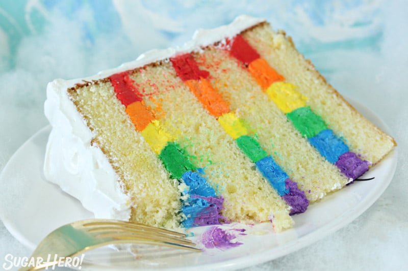 Close-up of rainbow cake slice on a white plate, with a bite taken out of it