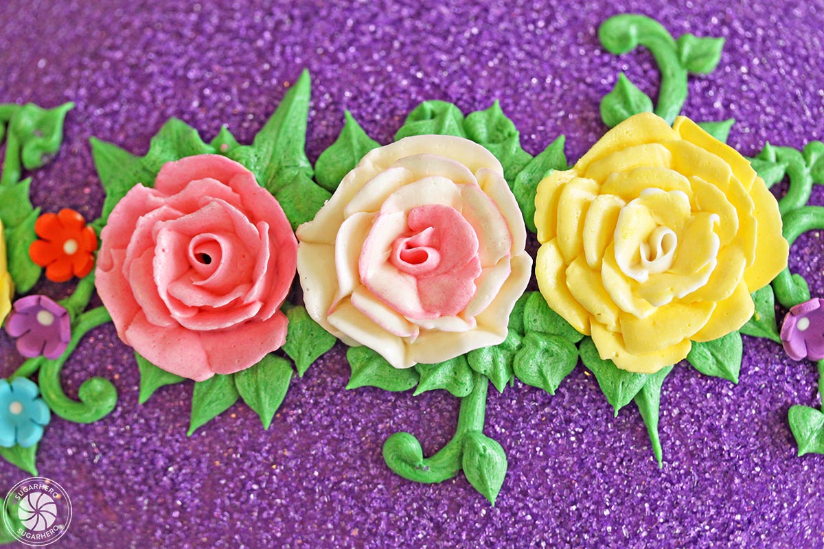 Three buttercream roses on a background of green leaves and purple sparkling sugar