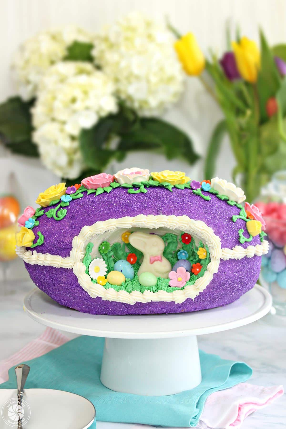 Looking for a knockout Easter cake? Try this Sugar Easter Egg Cake! It's based on old-fashioned panoramic sugar eggs, but it's made out of CAKE! It's entirely edible and the perfect Easter dessert! | From SugarHero.com Sugar Easter Egg Cake on a white cake stand in front of hydrangeas and tulips