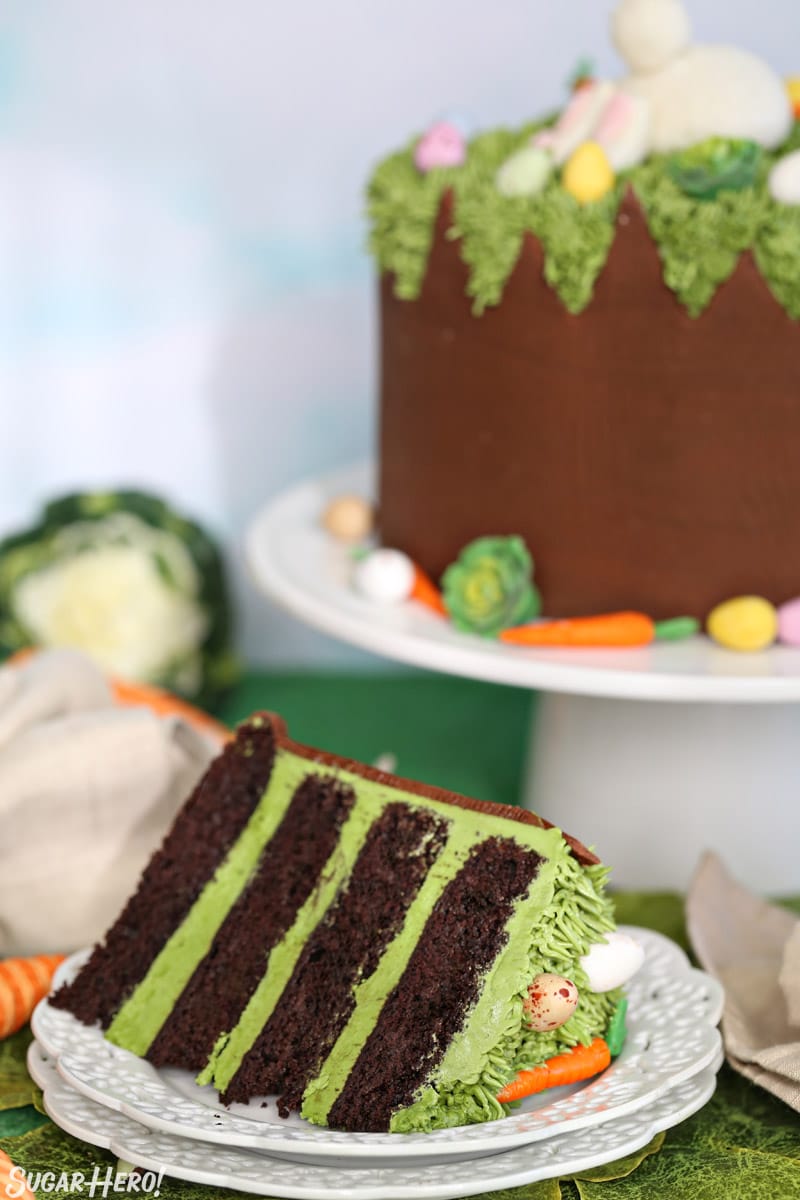 A slice of Chocolate Easter Bunny Cake on a white plate, with the whole cake in the background on a white cake stand