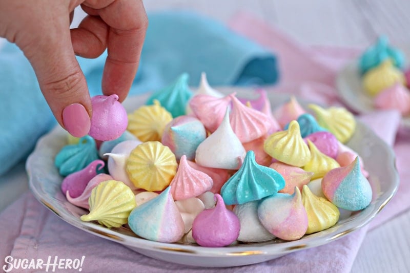 Hand picking up purple meringue kiss from plate of assorted meringues