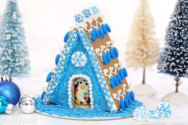 Blue-themed A-frame gingerbread house, with royal icing snowflakes and a royal icing penguin peeking out from inside