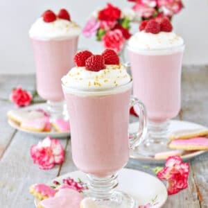 3 mugs of Raspberry White Hot Chocolate next to flowers and cookies.