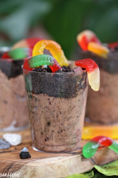 Clear plastic cup containing chocolate pudding, Oreo crumbs, and gummy worms, on a wooden cutting board.