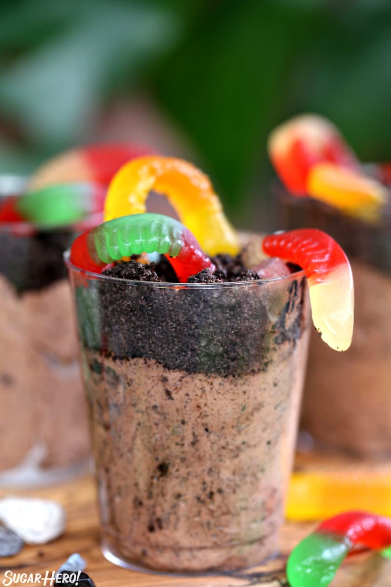 Chocolate pudding cup with oreo crumbs and gummy worms, on a wooden board.