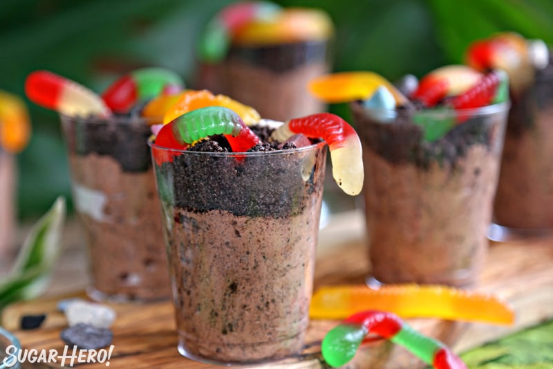Group of dirt pudding cups on wooden board wiht gummy worms scattered around.