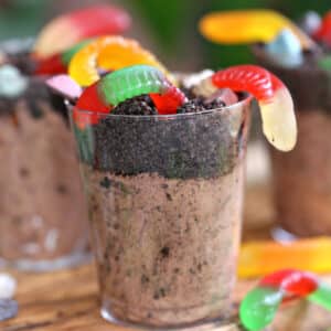 Clear plastic cup containing chocolate pudding, Oreo crumbs, and gummy worms, on a wooden cutting board.