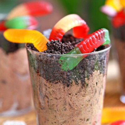 Clear plastic cup containing chocolate pudding, Oreo crumbs, and gummy worms, on a wooden cutting board.