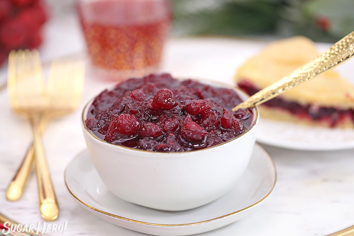 Homemade cranberry sauce in a white bowl with a gold spoon and cranberry cake in the background.
