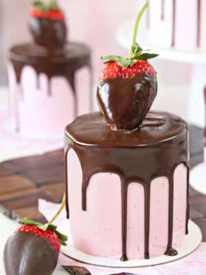 Close-up of mini cake with pink strawberry buttercream, a chocolate drip, and a large chocolate strawberry on top.