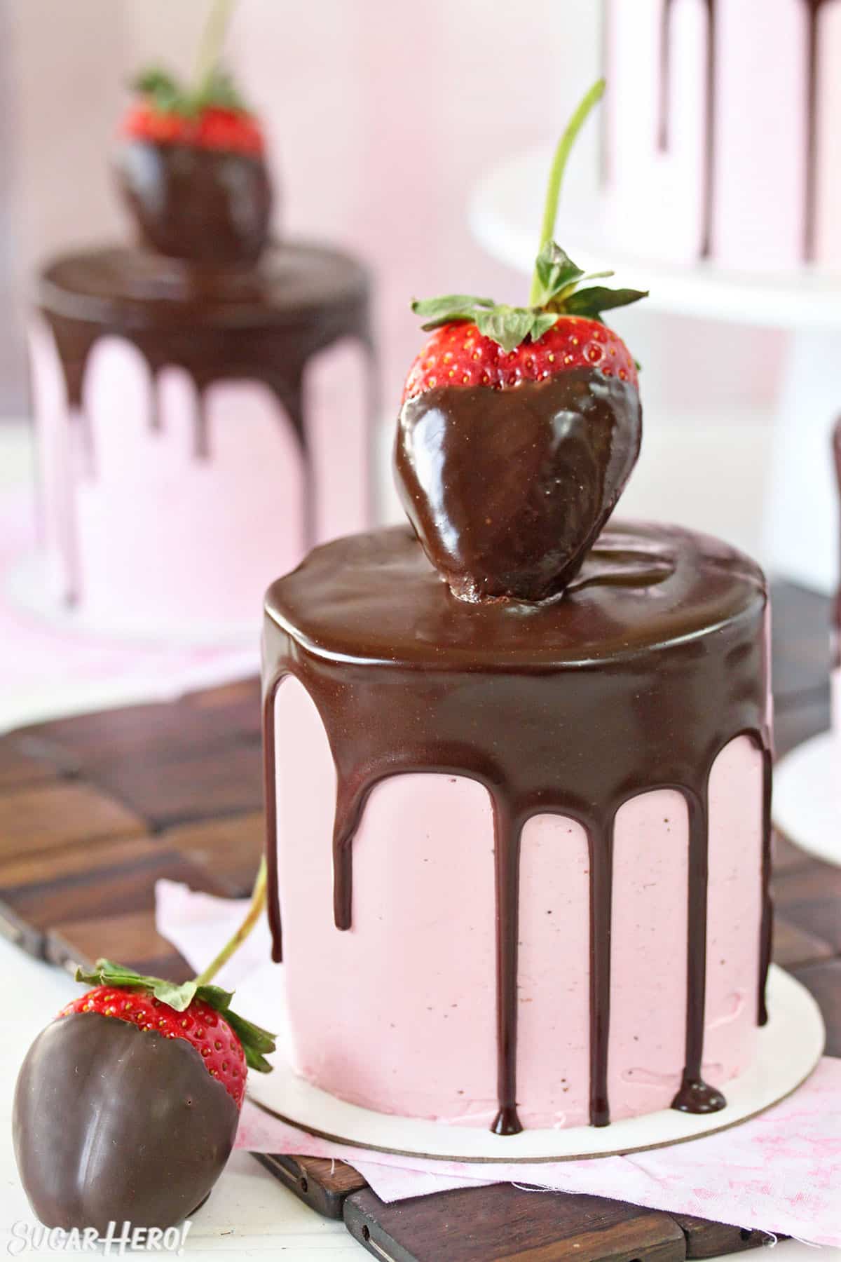 Close-up of mini cake with pink strawberry buttercream, a chocolate drip, and a large chocolate strawberry on top.