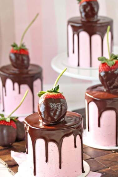 Four mini chocolate cakes, covered with strawberry buttercream, a chocolate drip, and a chocolate-dipped strawberry on top.