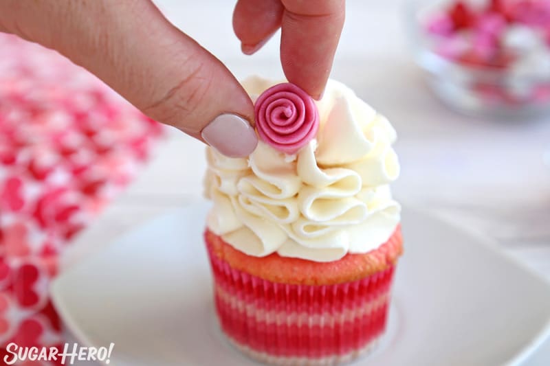 Hand placing a fondant ribbon rose on a ruffled white cupcake