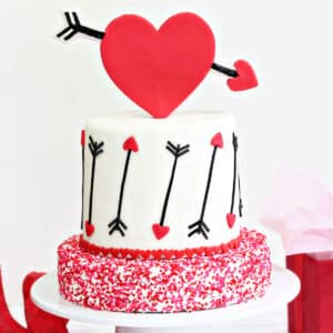 A Red Velvet Valentine's Day Cake on a white cake platter next to a gift box and ribbon.