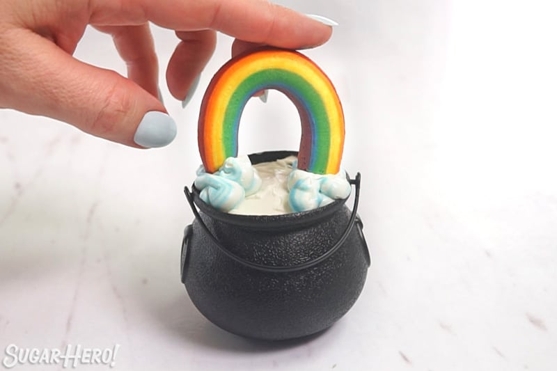 A hand placing a buttercream rainbow on top of a cupcake pot