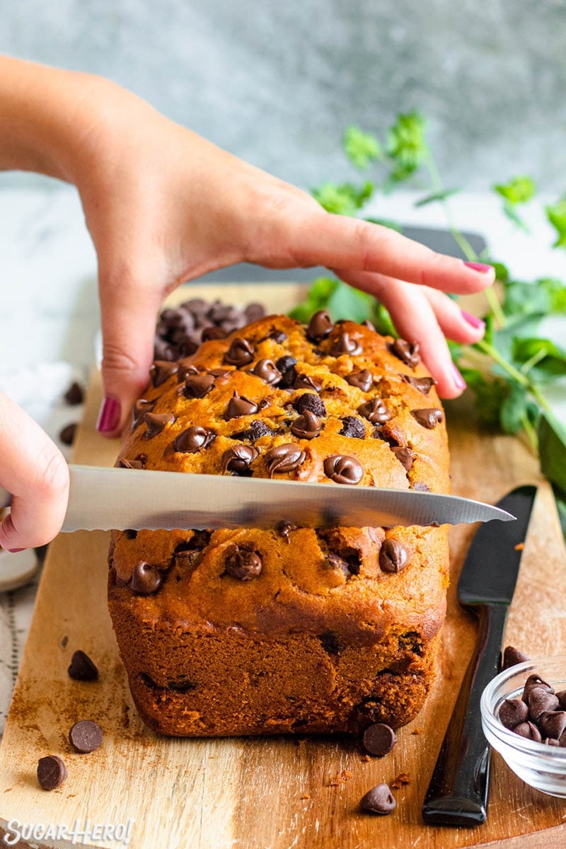 Hands slicing a loaf of pumpkin chocolate chip bread on a wooden cutting board.