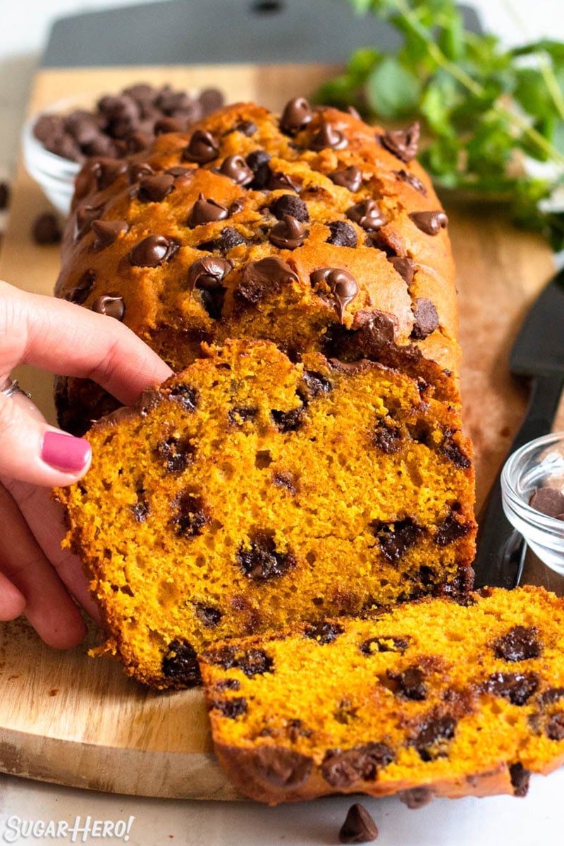 Hand removing a slice of pumpkin chocolate chip bread from the rest of the loaf on a wooden cutting board.