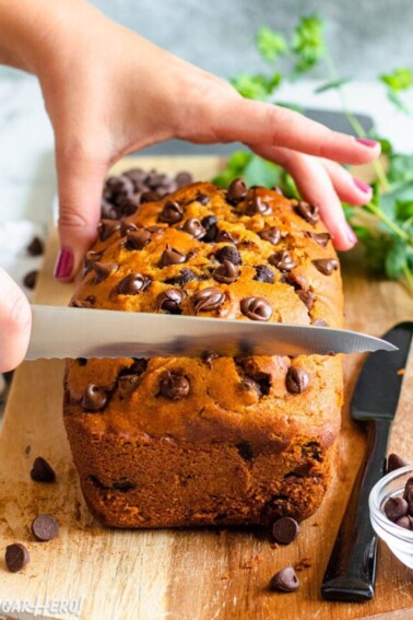 Hands slicing a loaf of pumpkin chocolate chip bread on a wooden cutting board.