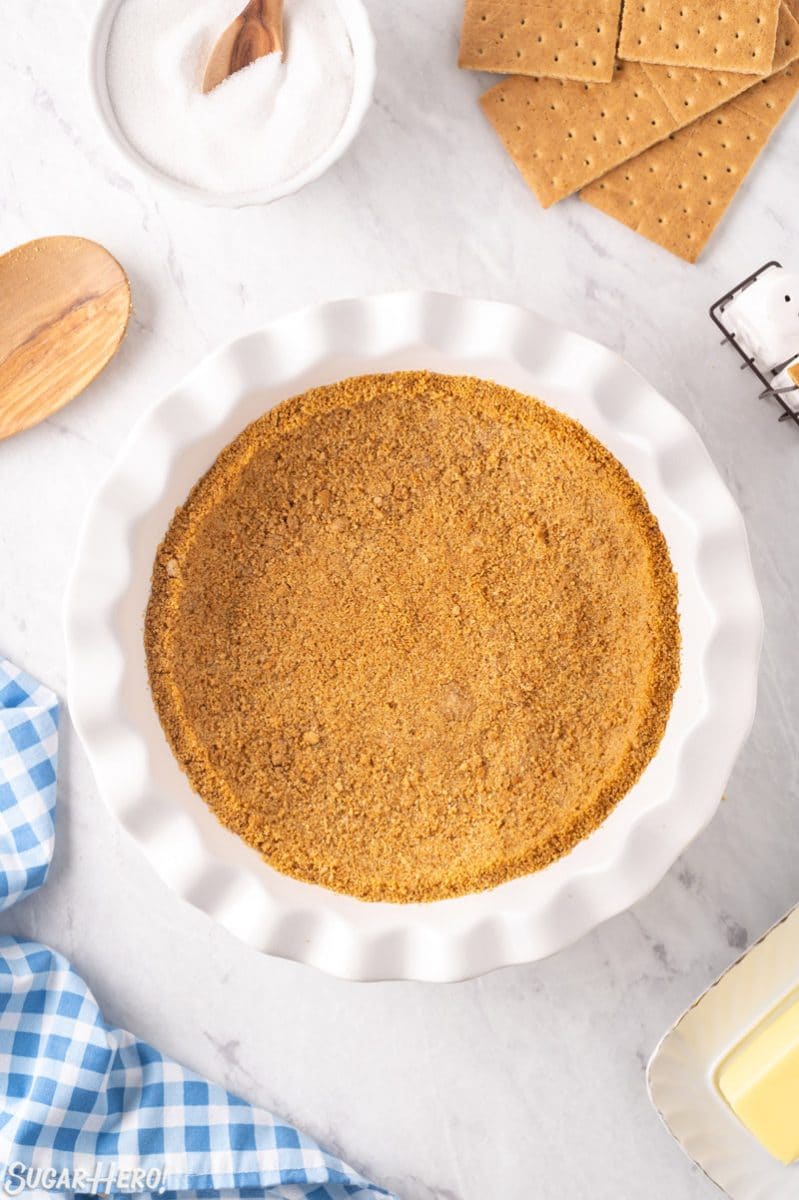 Photo of graham cracker crust in a white pie pan with ingredients surrounding it.