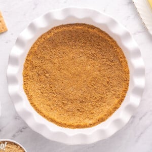 Photo of graham cracker crust in a white pie pan with ingredients surrounding it.