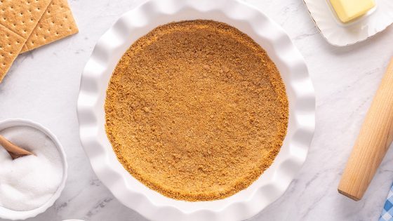 Photo of graham cracker crust in a white pie pan with ingredients surrounding it.