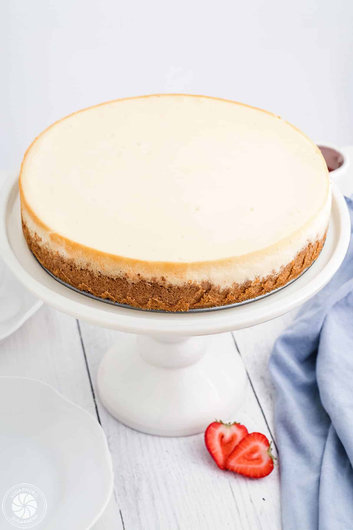 Plain cheesecake on a white cake stand with sliced strawberries and a blue napkin at the base.