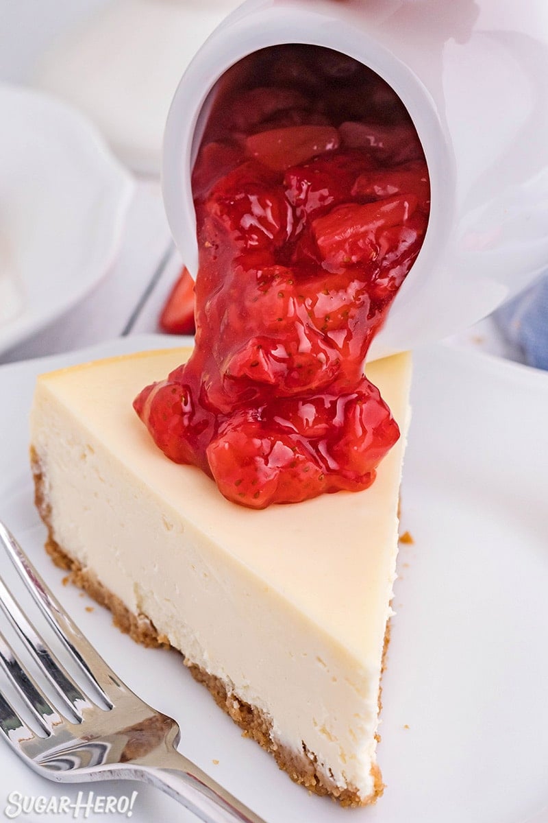 Pouring strawberry sauce from a pitcher onto a slice of cheesecake.