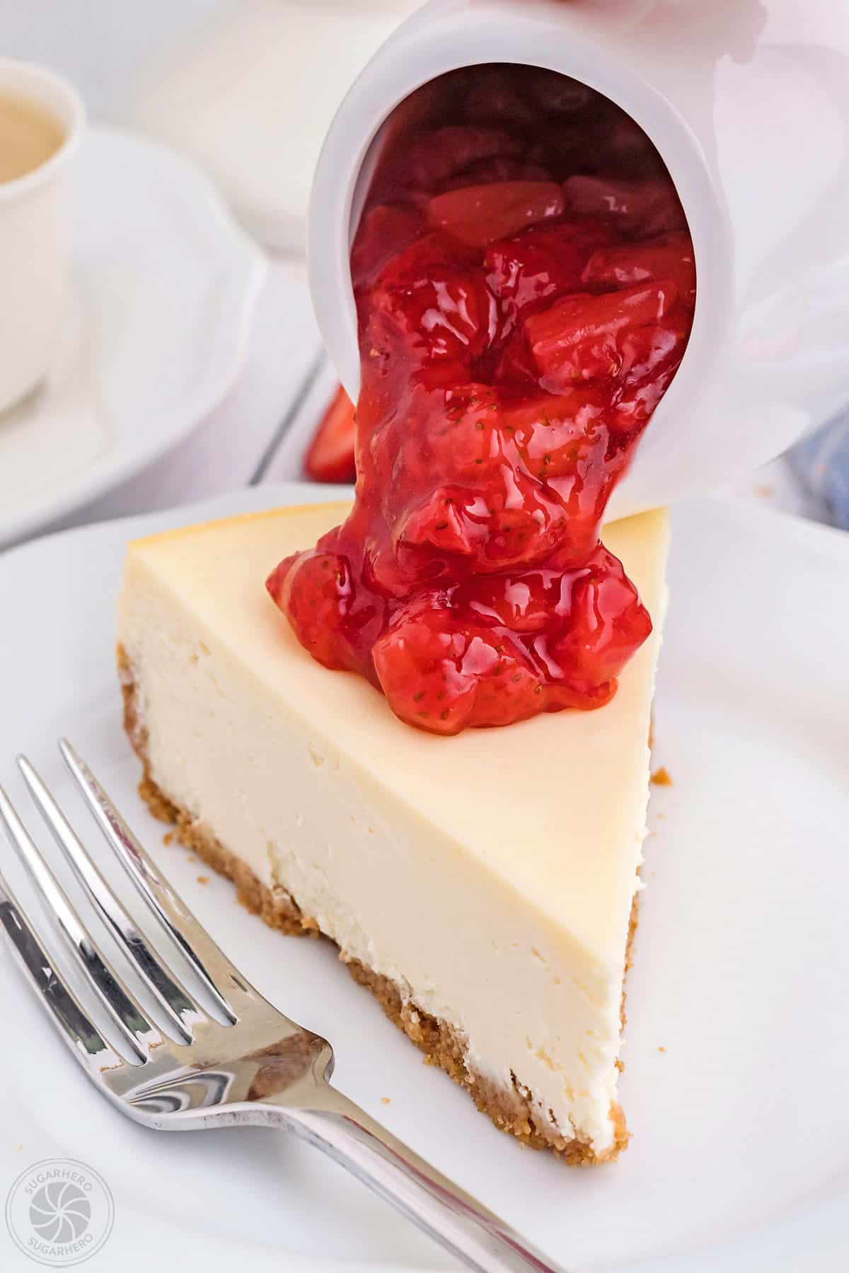 Pouring strawberry sauce from a pitcher onto a slice of cheesecake.