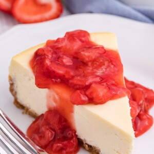 New York Style Cheesecake slice on a white plate with strawberry sauce dripping off the top.