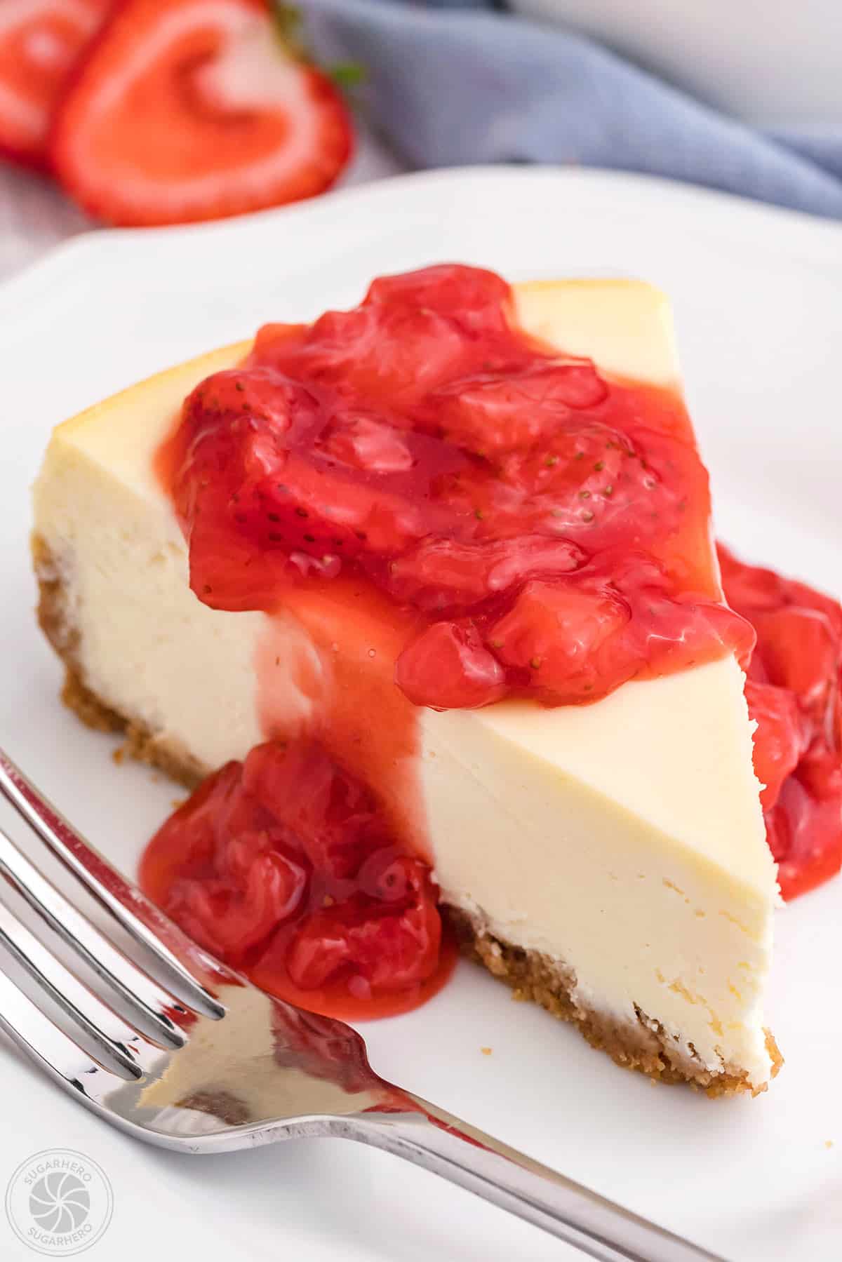 New York Style Cheesecake slice on a white plate with strawberry sauce dripping off the top.