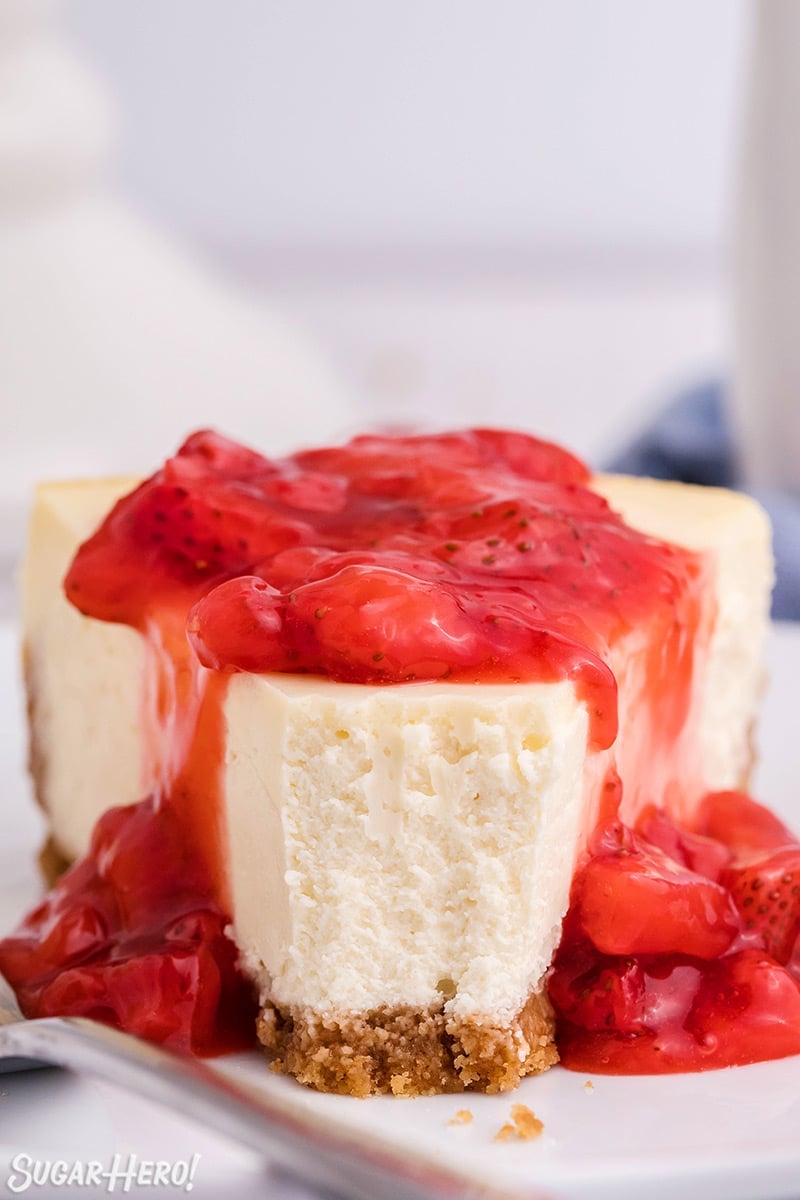 Close-up slice of white cheesecake with strawberry sauce on top and a bite taken out of it.